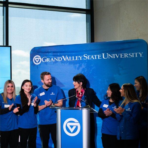 nursing students stand on either side of President Mantella, podium with GV on front and banner with GVSU spelled out in back