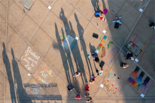  As seen from a drone, shadows of people are cast over colorful chalk art drawings on a sidewalk. 