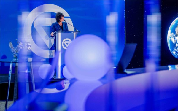  Among blue lights, a college president speaks at a lectern. 