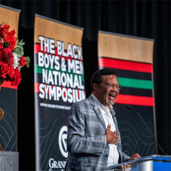 Greg Mathis in plaid suit jacket stands at podium, behind him black, red, green and brown banner The Black Boys and Men National Symposium