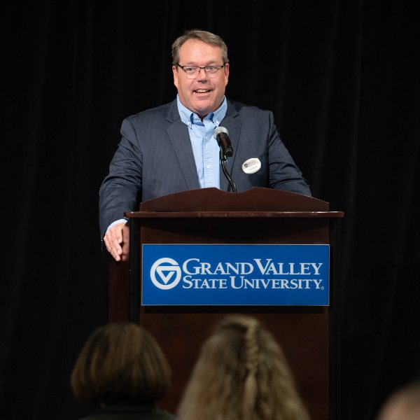 Greg Warsen at podium in blue suit coat and light blue shirt with no tie; podium has GVSU wordmark on it